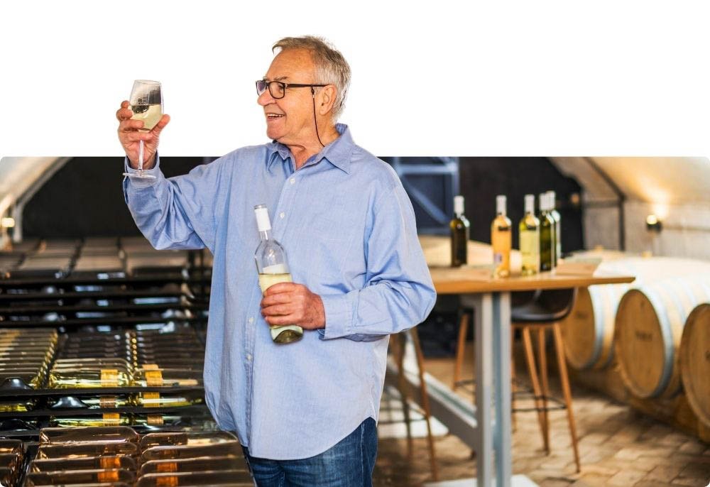 A imagem mostra um homem mais velho em uma vinícola, segurando uma garrafa de vinho branco em uma das mãos e um copo de vinho na outra. Ele parece estar apreciando o vinho, olhando para o copo com um sorriso. Ao fundo, há garrafas de vinho em uma mesa e barris de vinho ao longo das paredes, indicando que ele está em uma adega ou vinícola. O ambiente é iluminado e aconchegante, com um toque rústico.